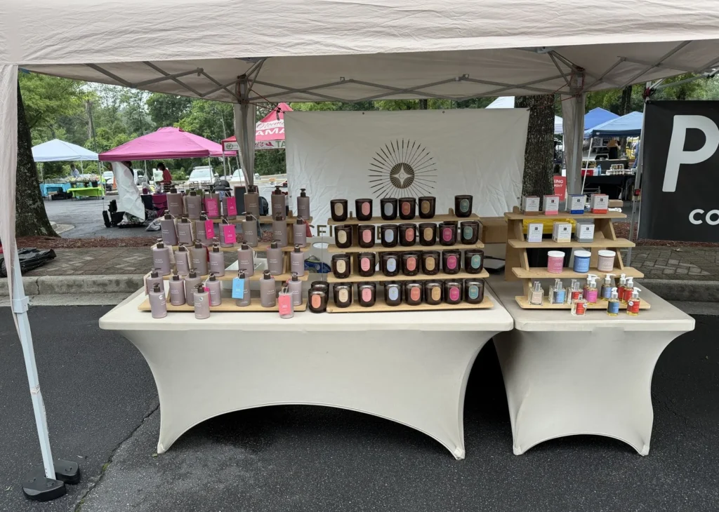Celeste Naturals' farmers market booth today, tiered wooden risers of candles and lotions under a branded backdrop.